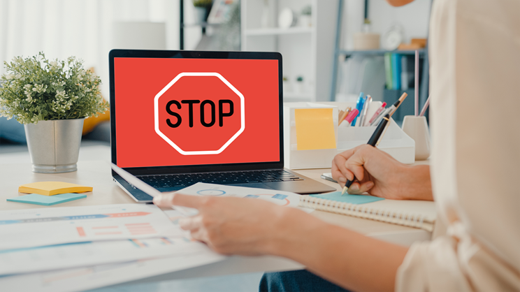 A person writes in a notebook at a desk with charts and papers, while their work computer displays a large red stop sign on the screen.