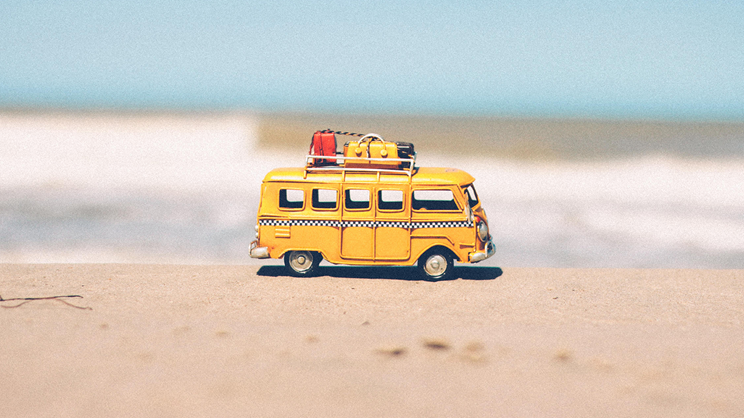 A small yellow toy van in work mode, with luggage on its roof, sits on sandy ground at a beach, with blurred ocean waves and sky in the background.