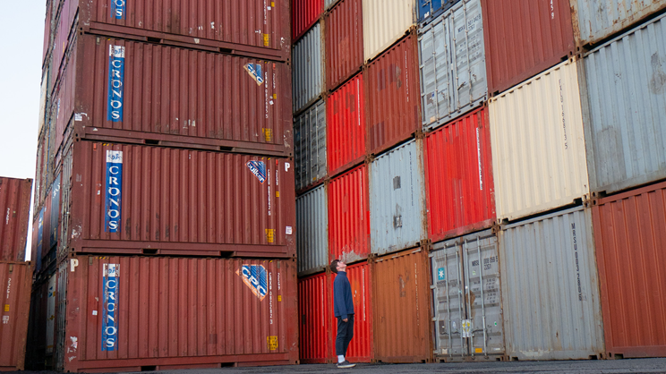 A person in a blue jacket and jeans stands in front of tall stacks of colorful shipping containers, looking up at the impressive storage arrangement.