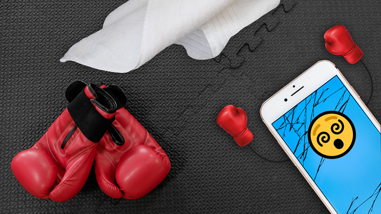 A pair of red boxing gloves, a crumpled white cleaning wipe, and a cracked tech smartphone with a dizzy face emoji on the screen lie on a textured black surface. The phone sports tiny red boxing gloves as earphones.