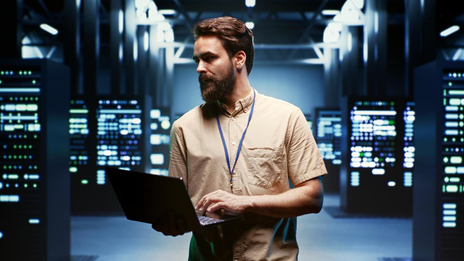 A man with a beard stands in a server room, holding and typing on a laptop. He wears a beige shirt and lanyard, surrounded by rows of illuminated server racks—an environment ideal for secure web hosting under bright overhead lights.